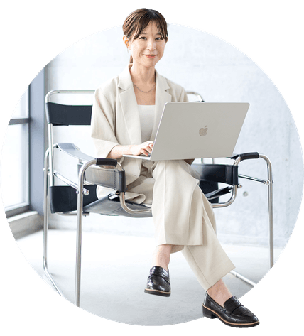 アイボリーのセットアップスーツで椅子に座って正面を向いているアッシー。膝の上でMacBook Proを開いている