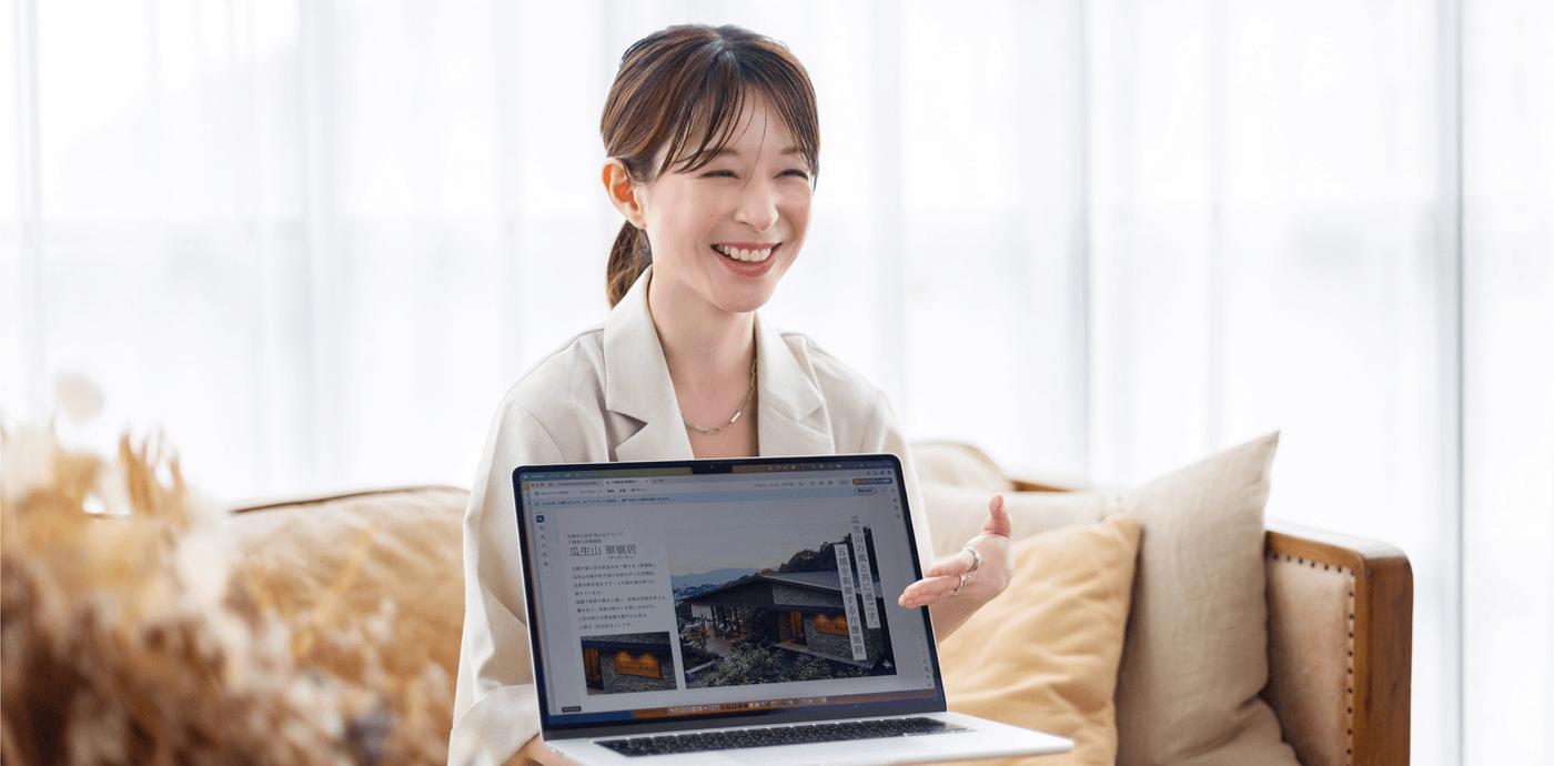 アイボリーのセットアップスーツで椅子に座り、MacBook Proの画面の正面に向け、笑顔で説明をしているアッシー
